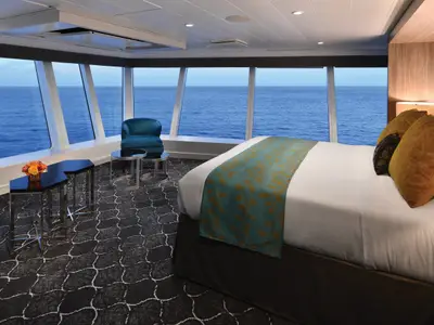 Blick in eine Kabine der Allure of the Seas mit raumhohen Fenstern und Meerblick. Sie zeigt ein Doppelbett, einen Sessel und Tische.