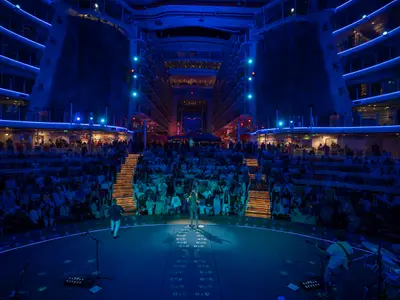 Eine Abendveranstaltung im AquaTheater der Allure of the Seas. Musiker treten auf einer Bühne auf, umgeben von einem sitzenden Publikum auf terrassierten Stufen und den beleuchteten Decks des Schiffes.