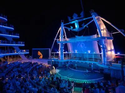 Die Allure of the Seas bei Nacht. Ein Live-Konzert auf der beleuchteten Bühne, mit einer Künstlerin auf dem Bildschirm, Publikum und blauen Lichtern.