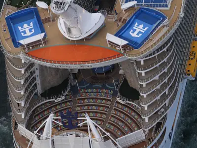Vogelperspektive auf die Allure of the Seas. Das offene AquaTheater mit Sitzreihen und Pool, umgeben von Balkonkabinen. Blaue Sportfelder mit Royal Caribbean Logo auf dem Oberdeck sichtbar.