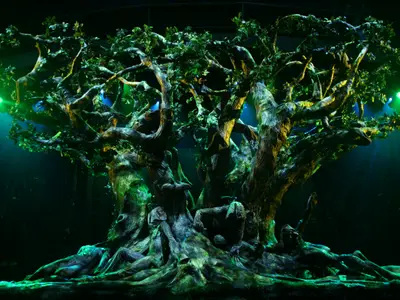 Nahaufnahme einer detailreichen Bühnenkulisse, die einen alten, majestätischen Baum darstellt. Grüne und blaue Scheinwerfer beleuchten die verdrehten Äste und Wurzeln, in denen sich versteckte Gesichter und Figuren befinden. Die Szene ist dunkel und atmosphärisch, vermutlich während einer Theateraufführung an Bord.