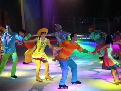 Eiskunstläufer in bunten Kostümen performen auf der Eisbahn der Allure of the Seas. Eine Gruppe hält sich an den Händen und gleitet über das von farbigen Lichtern erleuchtete Eis. Eine dynamische Show an Bord.