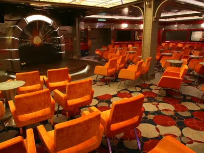 Ein Veranstaltungsraum auf der Allure of the Seas mit einer runden Holzbühne und einer Leinwand mit stilisiertem Sonnenmotiv. Zahlreiche orangefarbene Sessel mit kleinen Tischen sind in Reihen angeordnet, der Boden ist mit einem gemusterten Teppich ausgelegt.
