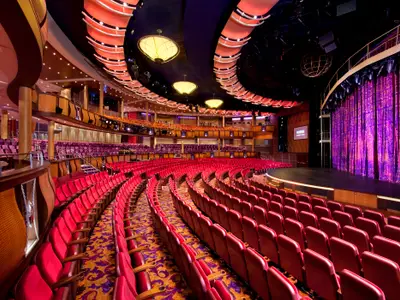 Das leere Haupttheater auf der Allure of the Seas. Reihen roter Polstersitze führen zu einer Bühne mit glitzerndem violettem Vorhang. Die geschwungene Decke ist beleuchtet.