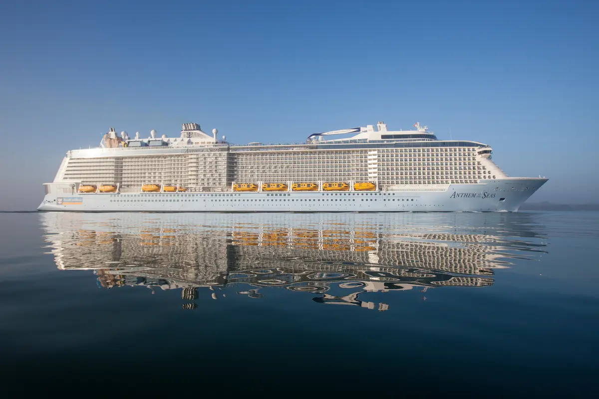 Die große weiße Anthem of the Seas gleitet auf spiegelndem, blauem Meer unter klarem Himmel. Gelbe Rettungsboote und zahlreiche Balkone sind an der Schiffswand zu erkennen.