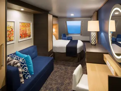 Geräumige Kabine auf der Anthem of the Seas mit blauem Sofa, Doppelbett, Schreibtisch und großem Fenster zum Meer. Ein beleuchteter Spiegel und Kunstwerke schmücken den Raum.