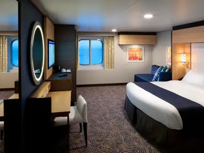 Kabinenansicht auf der Anthem of the Seas: Ein Bett, blaues Sofa und Schreibtisch mit TV sind zu sehen. Zwei Bullaugen bieten Meerblick, der Boden hat einen gemusterten Teppich.