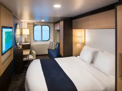 Pressefoto einer modernen Kabine der Anthem of the Seas. Zu sehen sind ein Doppelbett, Schreibtisch, Sofa am ovalen Fenster und ein Fernseher. Einrichtung in Holz, Weiß, Blau.