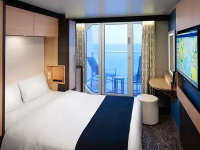 Eine moderne Balkonkabine an Bord der Anthem of the Seas. Zu sehen sind ein gemachtes Bett, ein Balkon mit Meerblick und Stühlen, sowie ein Bereich mit TV und einem kleinen Tisch.