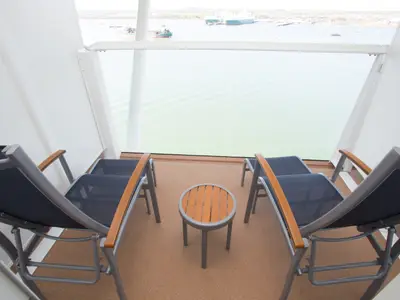 Ansicht des privaten Balkons an Bord der Anthem of the Seas. Zwei Liegestühle und ein Beistelltisch stehen bereit. Im Hintergrund ist ein Hafen mit Schiffen und Land zu sehen.