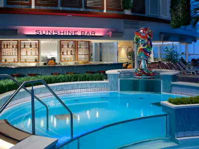Innenpoolbereich auf der Anthem of the Seas: Blauer Pool, "Sunshine Bar" mit Flaschen, farbige Skulptur. Liegestühle im Hintergrund.