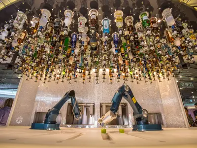 Eine futuristische Bar an Bord der Anthem of the Seas mit hunderten von Spirituosenflaschen, die von der Decke hängen. Zwei Roboterarme bereiten Getränke auf dem Tresen zu, einer hält ein grünes Getränk.