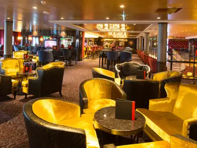 Lounge der Anthem of the Seas. Gold-gelbe Samtsessel, dunkle Tische und eine Bar dominieren den Raum. Ein roter Bühnenbereich im Hintergrund. Luxuriöses Interieur.