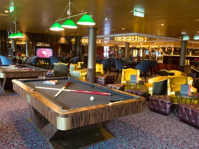 Die elegante Lounge der Anthem of the Seas zeigt zwei Billardtische mit grünen Lampen. Viele gelbe Sessel, braune Lederhocker und eine Bar im Hintergrund prägen den Raum.