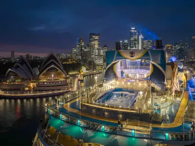 Das Heck der Anthem of the Seas bei Nacht, mit beleuchtetem FlowRider-Surfsimulator, Kletterwand und dem ikonischen Opernhaus Sydney sowie der Skyline der Stadt im Hintergrund. Ein Passagier surft auf dem FlowRider.