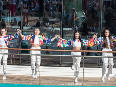 Vier Tänzerinnen in Flaggen-Jacken und weißen Outfits springen auf dem Deck der Anthem of the Seas. Sie halten Händchen, im Hintergrund Passagiere in der Glasfassaden-Spiegelung.