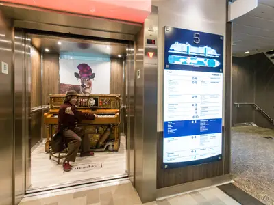 Ein Musiker spielt Piano im kunstvoll dekorierten Aufzug der Anthem of the Seas, Etage 5. Rechts ein beleuchteter Deckplan.