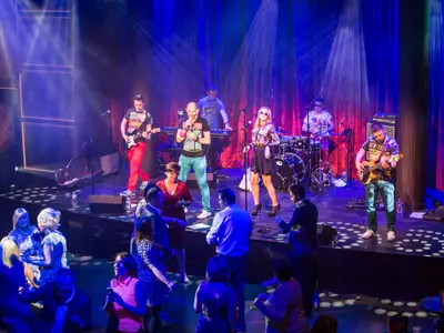 Eine Live-Band spielt auf einer Bühne mit blauen Lichtern und roten Vorhängen. Gäste tanzen und beobachten die Aufführung. Sängerin und Musiker sind zu sehen.