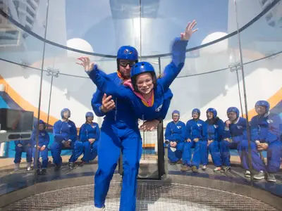 Indoor-Skydiving-Erlebnis auf der Anthem of the Seas. Ein Instruktor hilft einer lächelnden Frau im Windkanal zu schweben, während andere Teilnehmer zuschauen.
