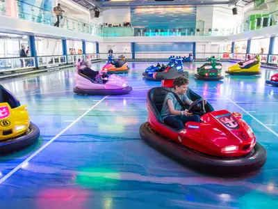 Bumper-Car-Arena an Bord der Anthem of the Seas. Gäste in bunten Autos auf dem Boden. Ein lächelnder Junge im roten Wagen ist im Vordergrund. Zuschauer auf oberen Decks beobachten.