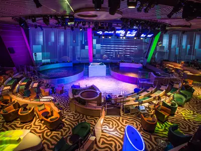 Innenansicht des Two70 Entertainment-Centers auf der Anthem of the Seas. Zentrale Bühne, Wasserbecken und viele Sitzbereiche in bunter Lichtstimmung.