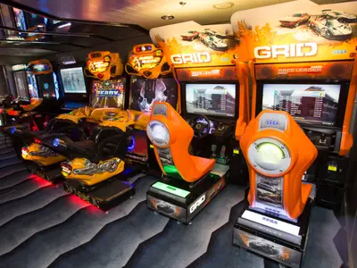 Arcade-Raum auf der Anthem of the Seas mit vielen Rennsimulatoren. Orangefarbene GRID- und gelbe Motorrad-Spiele mit leuchtenden Lichtern und Displays. Teppichboden mit Wellenmuster.