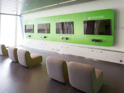 Der Gaming-Bereich der Anthem of the Seas zeigt vier Xbox One Konsolen an einer grünen Wand mit Bildschirmen. Bequeme Sessel in Grau und Grün stehen davor, links sind große Fenster.