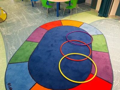 Spielbereich auf der Anthem of the Seas mit großem blauen Teppich, bunten Rändern und drei Hula-Hoops (gelb, rot, violett). Helle Böden und grüne Stühle im Hintergrund.