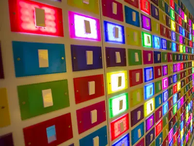 Nahaufnahme einer weißen Wand, die mit vielen rechteckigen, bunten Paneelen bedeckt ist. In jedem Paneel befindet sich ein weißer Lichtschalter. Einige Paneele leuchten hell in Farben wie Rot, Magenta, Blau, Gelb und Grün, während andere unbeleuchtet sind.