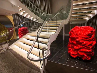 Geschwungene, beleuchtete Holztreppe mit Glasgeländer in einem Atrium der Anthem of the Seas. Zwei große, rote, skulpturale Kunstwerke flankieren die Treppe auf dunklem Boden.