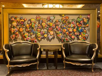 Zwei elegante, dunkle Ledersessel mit goldenen Verzierungen und pelzigen Armlehnen stehen vor einer großen Wandinstallation aus vielen bunten Totenkopf-Motiven in einem luxuriösen Raum.