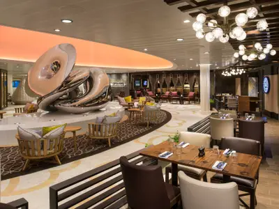 Eine moderne Lounge in der Anthem of the Seas mit einer großen, glänzenden Skulptur im Zentrum. Angrenzend sind ein Barbereich und mehrere Essbereiche mit gedeckten Tischen und Stühlen zu sehen.
