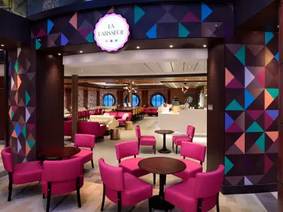 Blick in die La Patisserie der Anthem of the Seas. Der Raum ist gefüllt mit pinken Stühlen und Sofas, dunklen Tischen. Wände mit bunten geometrischen Mustern und runde Fenster zum Meer.
