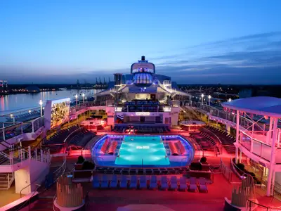 Blick auf das beleuchtete Pooldeck der Anthem of the Seas bei Dämmerung. Blauer Pool, rote Whirlpools, Liegestühle, Bar und Großbildschirm sichtbar. Im Hintergrund ein beleuchteter Hafen mit Kränen.