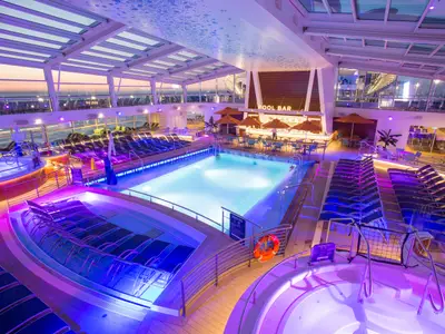 Der beleuchtete Innenpoolbereich der Anthem of the Seas am Abend zeigt einen großen Pool, Whirlpools, Liegestühle und eine Poolbar unter einem Glasdach mit Meerblick.