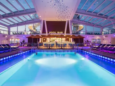 Innenpoolbereich der Anthem of the Seas mit beleuchtetem Pool, Poolbar, Liegestühlen und Glasdach. Violett-blaue Beleuchtung. Eine Person an der Bar.