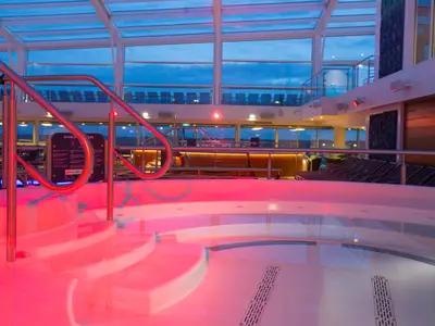 Abend im Solarium der Anthem of the Seas. Roter Whirlpool im Vordergrund. Panoramafenster mit Blick auf den blauen Himmel. Bar und Liegestühle im Hintergrund.