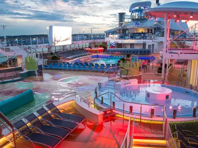 Abendliche Ansicht des Pooldecks der Anthem of the Seas. Mehrere beleuchtete Pools, ein Surfsimulator, Liegestühle und eine große Videoleinwand schaffen eine lebendige Atmosphäre.