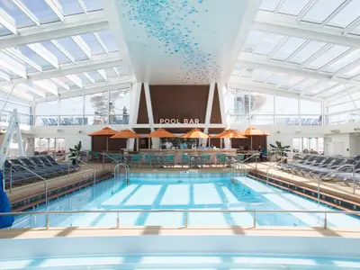 Innenpoolbereich der Anthem of the Seas mit großem Pool, einer Poolbar mit Sonnenschirmen und Liegestühlen unter Glasdach.