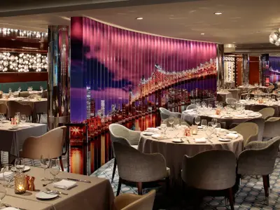 Elegantes Restaurant der Anthem of the Seas. Viele gedeckte Tische, dazu eine Wand mit Nachtpanorama der New Yorker Skyline und beleuchteter Brücke.