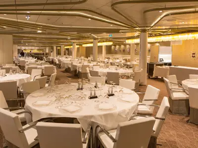 Ein eleganter, leerer Speisesaal auf der Anthem of the Seas. Viele Tische sind mit weißen Decken, Gläsern und Tellern gedeckt. Moderne weiße Stühle und eine warme Beleuchtung sind zu sehen.