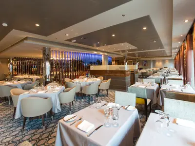 Restaurant auf Anthem of the Seas. Gedeckte Tische mit weißen Tischdecken, Gläsern, Besteck. Moderne Stühle, Teppich, Spiegelwand mit blauer Beleuchtung sind sichtbar.