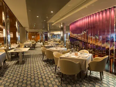 Innenansicht des stilvollen Restaurants auf der Anthem of the Seas. Viele gedeckte Tische, beigefarbene Stühle, gemusterter Teppich und eine leuchtende Stadtkulisse an der Wand.