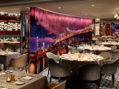 Das elegante Restaurant auf der Anthem of the Seas mit vielen eingedeckten Tischen. Eine Wand zeigt eine beleuchtete Stadt- und Brückenansicht bei Nacht.