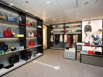 Store auf der Anthem of the Seas: Handtaschen, Schuhe, Kleidung in Regalen und an Stangen. Großes Poster mit Models, Armani Jeans sichtbar.