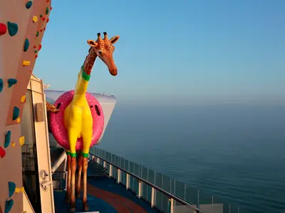 Eine gelbe Giraffenskulptur mit rosa Schwimmring steht auf dem Deck der Anthem of the Seas. Im Hintergrund ist das weite Meer zu sehen, links eine Kletterwand.