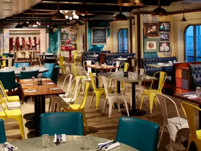 Jamie Oliver Restaurant auf der anthem of the Seas mit Tischen, gelben und petrolfarbenen Stühlen, dunklen Sitzbänken. Wände mit Kunst, Fenster mit Meerblick.
