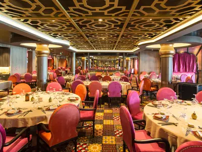 Das Hauptrestaurant auf der Anthem of the Seas. Viele festlich gedeckte Tische, umgeben von pinken und orangen Stühlen. Goldene Decke und bunter Boden.