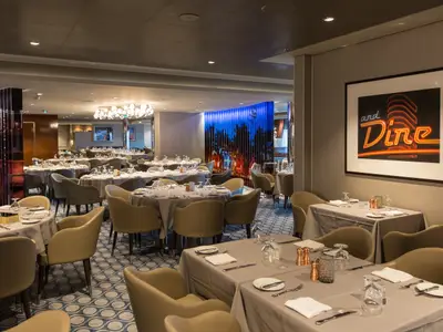 Modernes Restaurant der Anthem of the Seas mit zahlreichen gedeckten Tischen und beigen Stühlen. Auffällig sind die beleuchtete Wandkunst und ein Bild mit dem Neon-Schriftzug 'and Dine'.