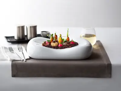 Eine kunstvoll angerichtete Gourmet-Speise mit Babykarotten und Wurzelgemüse in einem weißen Teller. Besteck und Weinglas daneben auf einem elegant gedeckten Tisch.